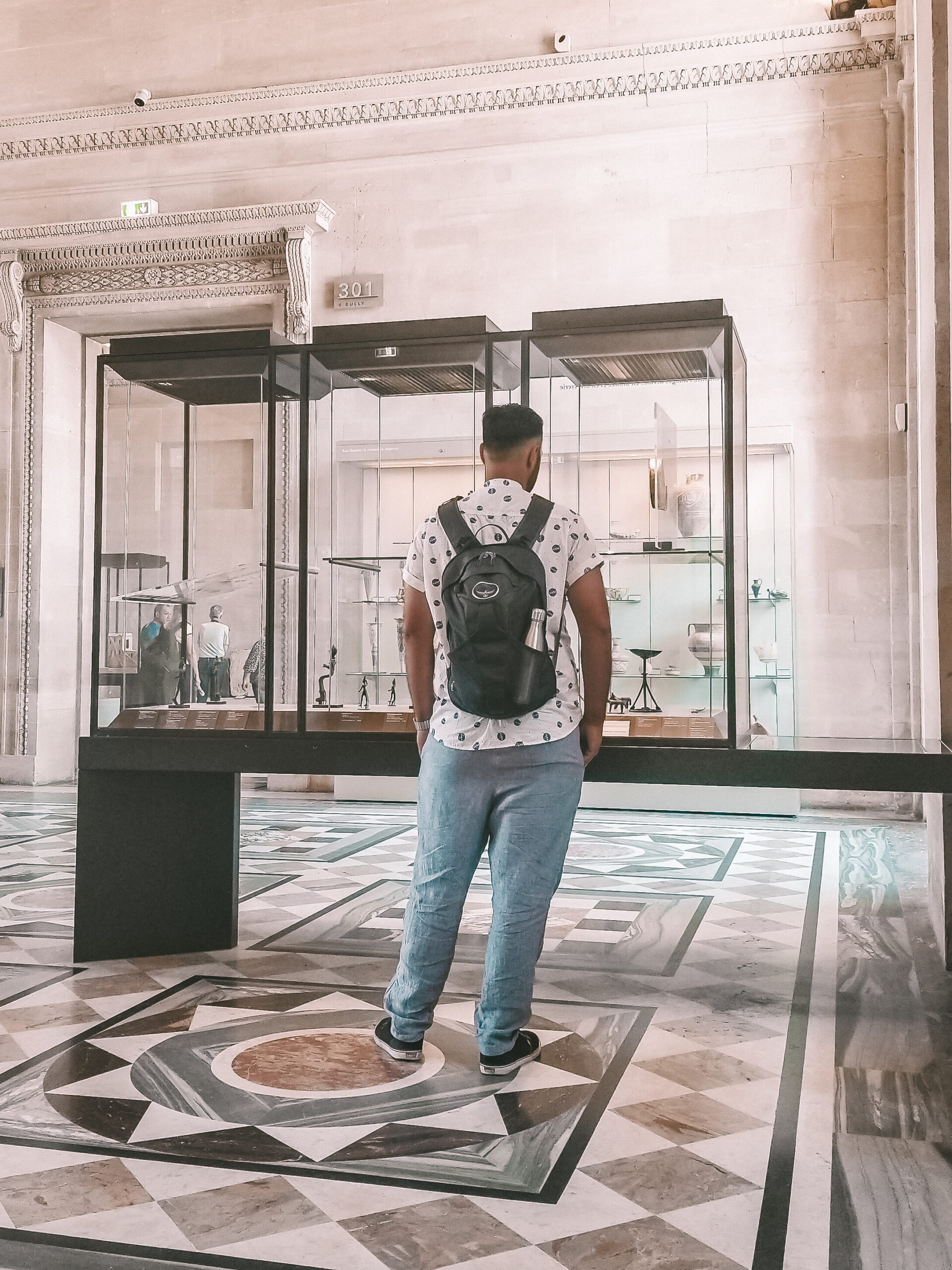 A man with a backpack viewing items in a museum. His back is turned to the viewer. 