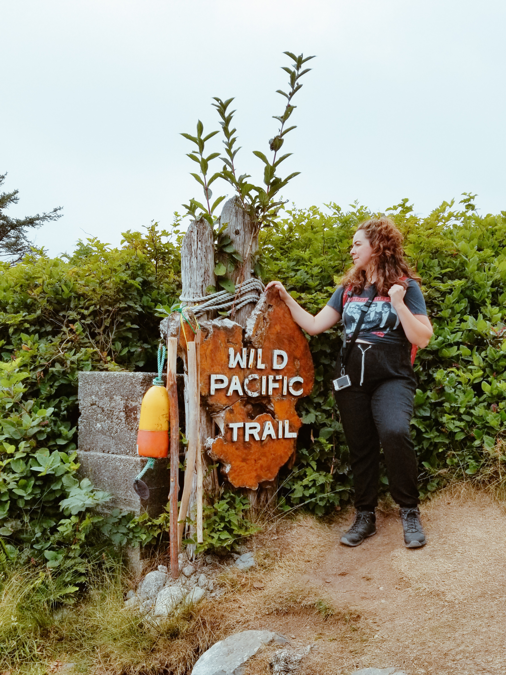 A woman with hiking boots and backpack leans against a sign that reads Wild Pacific Trail.