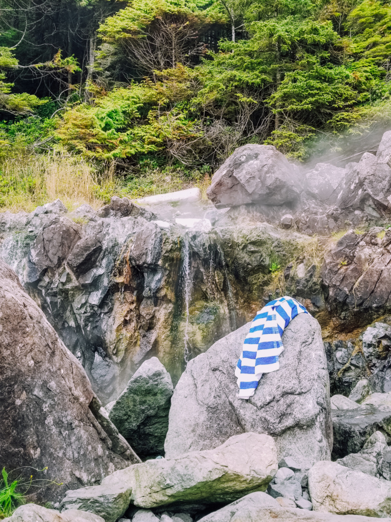 A blue and white striped towel is draped over a Rocks. Rocks are all around. In the center steam rises from a hot spring. 