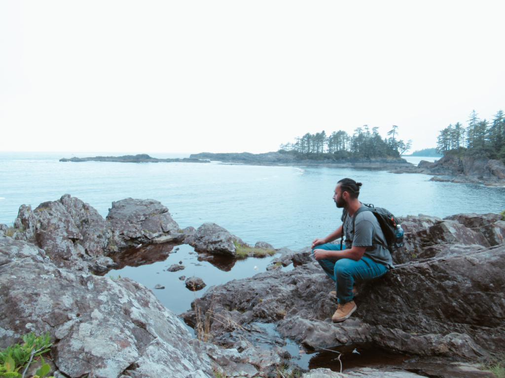 A man sits on a rock on a shoreline looking out to the Pacific Ocean. 